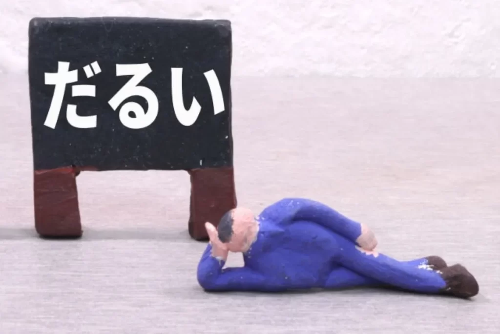 だるさで横になっている粘土人形の男性と、後ろに「だるい」と書かれた黒い看板のイメージ
