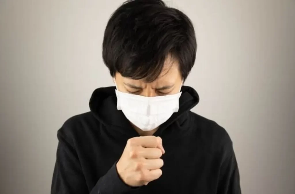 黒いパーカーを着た男性が頭を下げ、右手を口元に当てて咳をしている様子。喋ると咳が出る症状のイメージ。