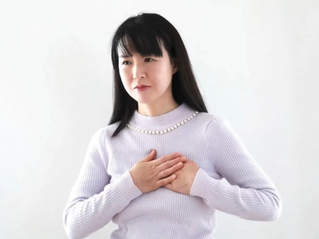 薄い紫色のセーターを着た女性が、胸を両手で押さえて痛みを感じている様子