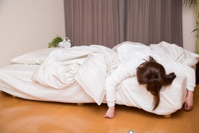 白い服を着た女性が白いベッドでうつ伏せに寝ており、枕の反対側で寝相が悪い様子
