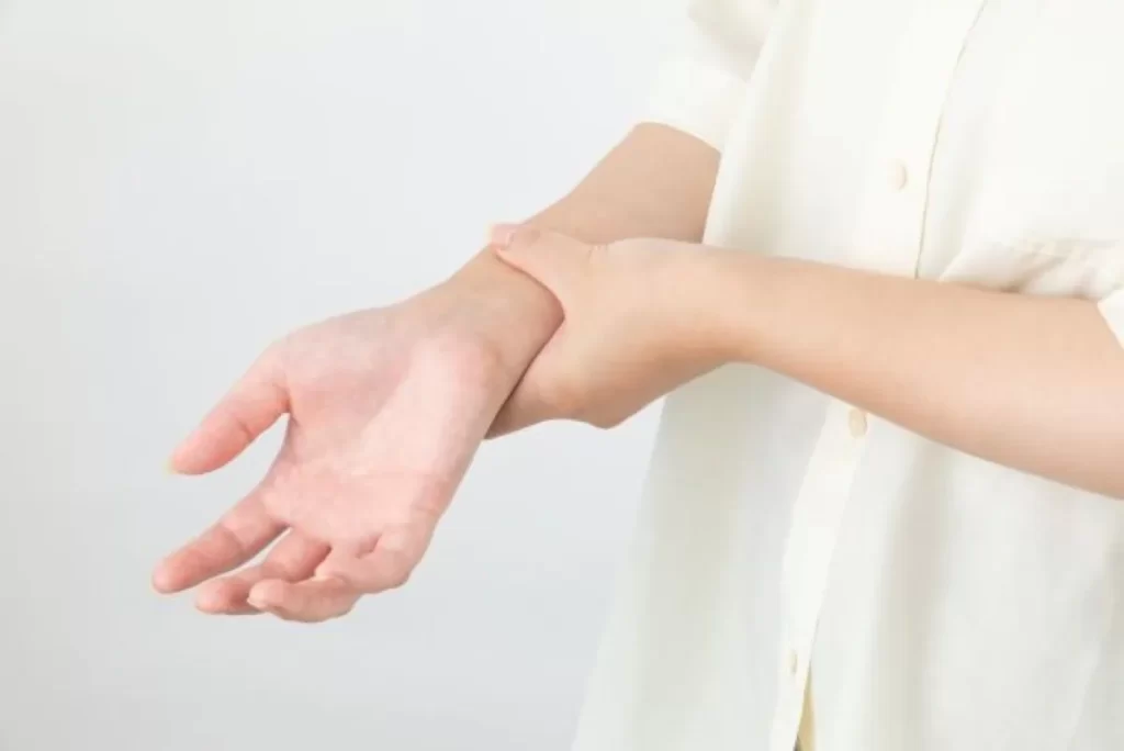 右手首の痛みを気にして左手で触っている女性（ベージュ半袖）のクローズアップ