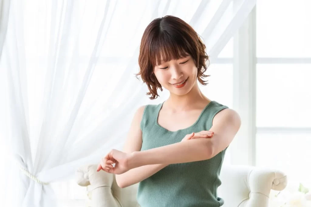 白いソファの前で、黄緑色のタンクトップを着た女性が腕の肌の改善を喜んで触っている様子。