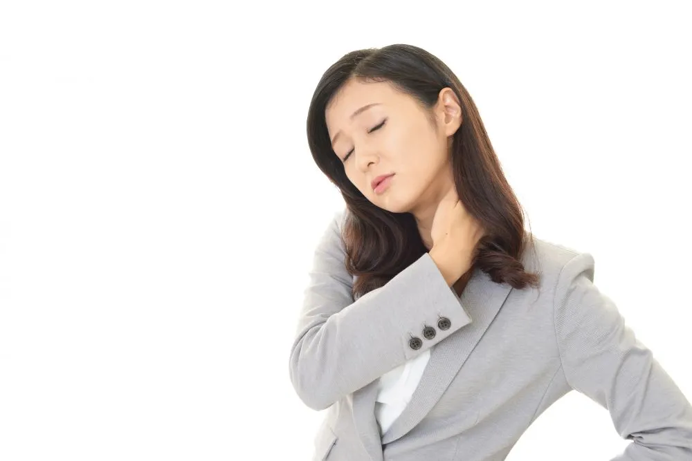 スーツ姿の女性が左首と肩の痛みに耐え触るクローズアップ画像。寝違いによる首肩痛の典型的な症状例。