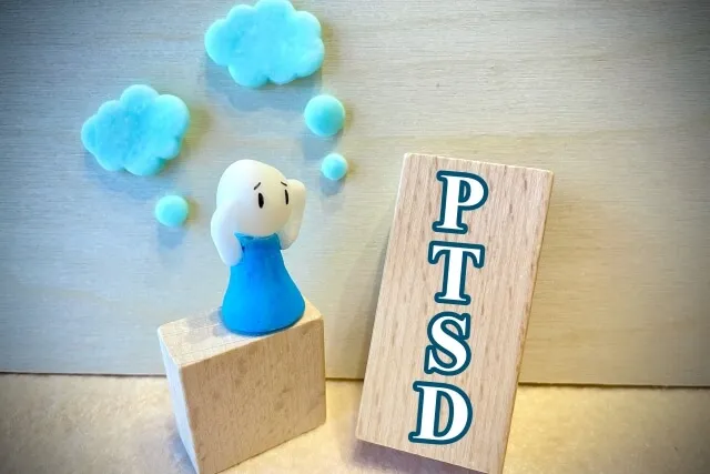 PTSDの文字が書かれた木の板の前で困った表情の人形が立つアイキャッチ画像。心的外傷後ストレス障害の症状や治療を象徴。