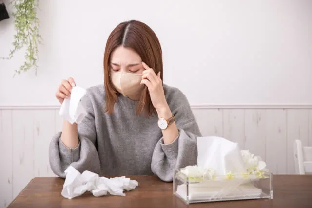 花粉症に悩む30代女性がグレーセーターとマスク姿で座り、テーブルにティッシュ箱を置き、使い捨てティッシュが山積みになった様子。鼻水症状の苦しみを象徴。