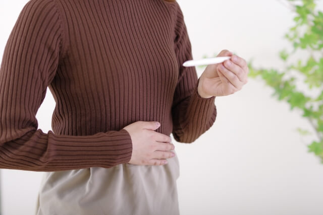 オックスブラッド色のセーターを着た女性が妊娠検査薬を持ちながらお腹を触る姿