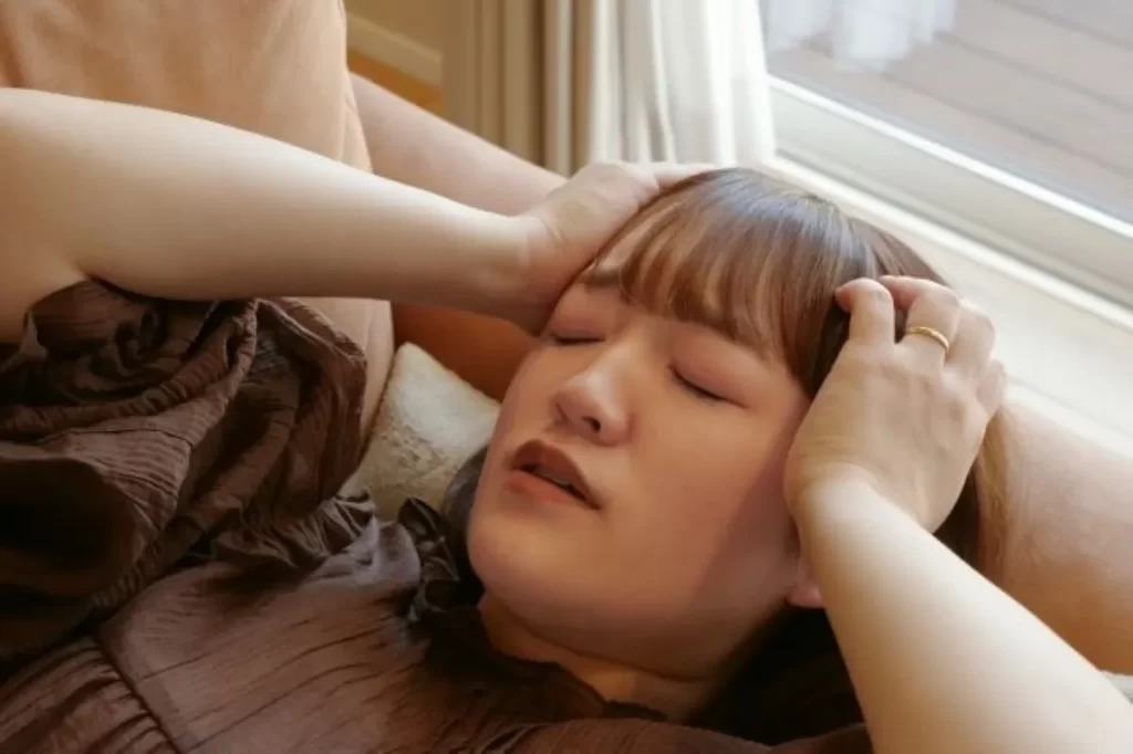 原因不明の激しいめまいでこめかみを押さえて苦しむ女性。ベッドから起き上がれない状態をイメージ