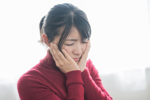 ワインレッドのセーターを着た女性が顔を両手で抑えている様子。顔面痙攣や鼻炎の症状をイメージ。