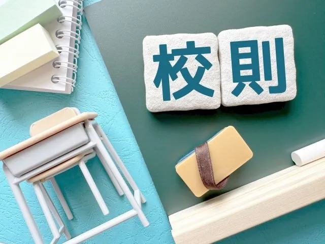 教室の机、椅子、メモ帳、緑の黒板と黒板消しが描かれたイラスト。黒板に「校則」の文字。