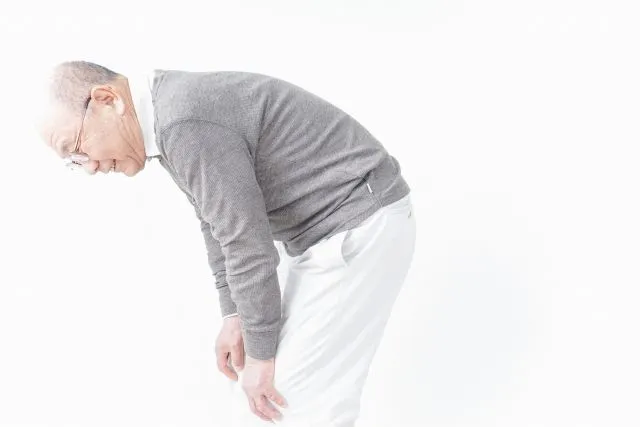 白いズボンとグレーのセーターを着た高齢男性が膝痛で前かがみになり膝を触っている様子。