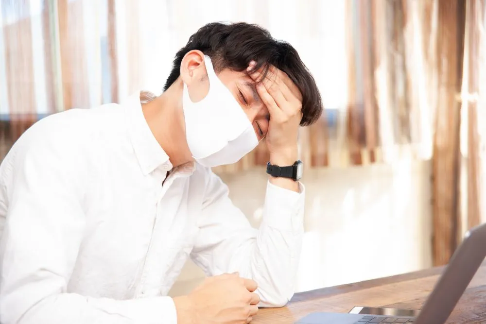 白いワイシャツとマスクを着用した男性が椅子に座り、発熱の痛みで左手で頭を触る様子