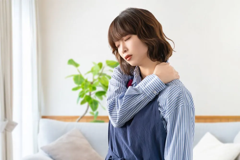 チェック柄のシャツを着た女性が左肩を触っている様子。首肩の痛みを表現。