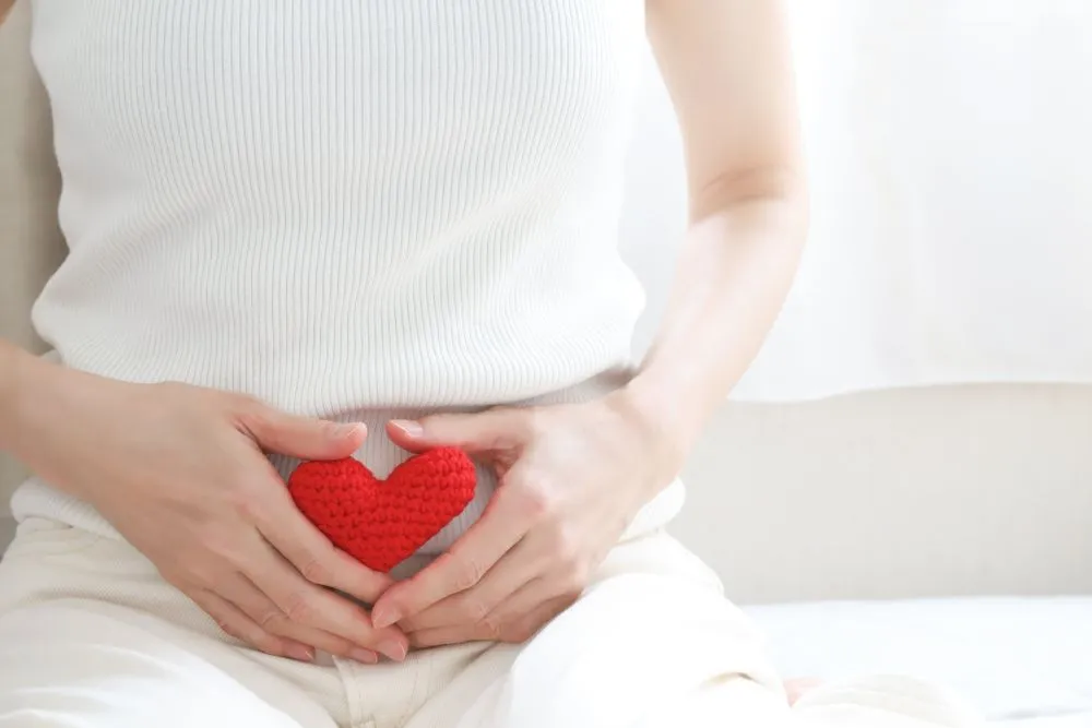 白い服を着た女性が座り、両手で赤いハートをお腹に持つイラスト。生理痛を和らげるツボの鍼治療をイメージ。