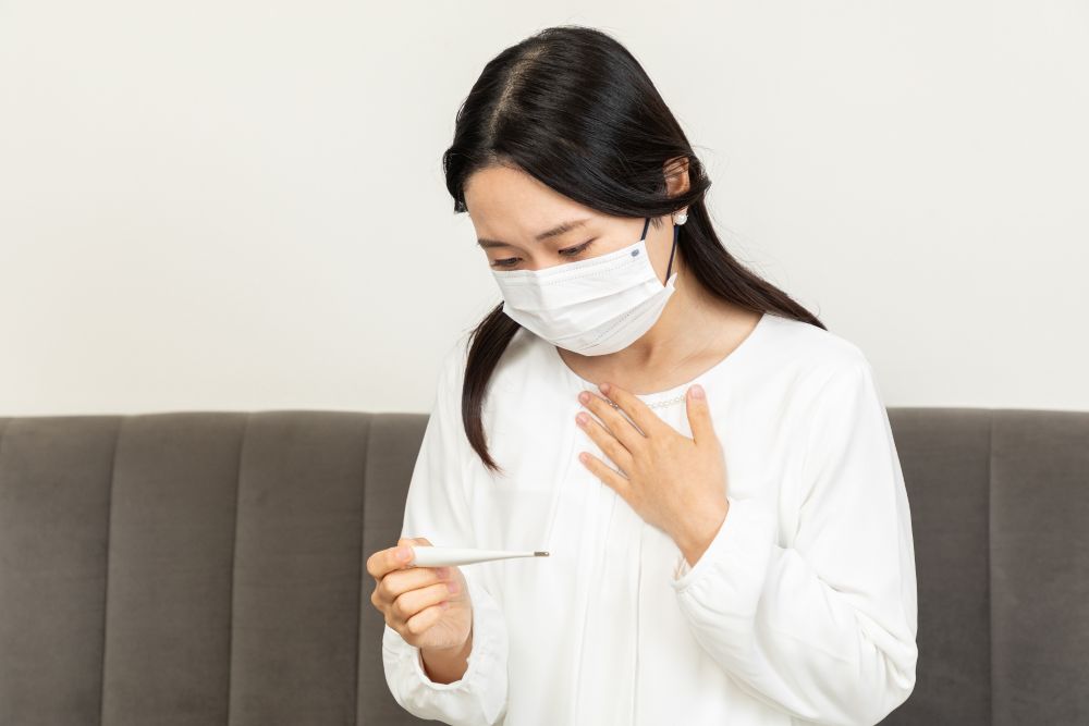 白い服を着たマスク姿の女性が温度計を持ち、心配そうに体温をチェックしている様子