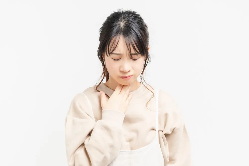 薄いグレー色の長袖シャツとエプロンを着た女性が、喉のあたりを気にするように触っている様子。バセドウ病の症状を表すイラスト。