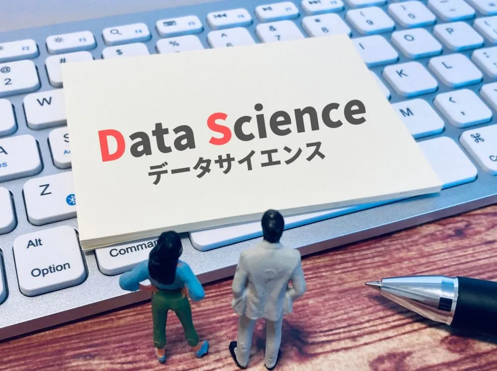 小さな男女人形の前に「データサイエンス」と書かれた看板。看板下にパソコンのキーボードとボールペンの一部が配置されたイラスト風画像。