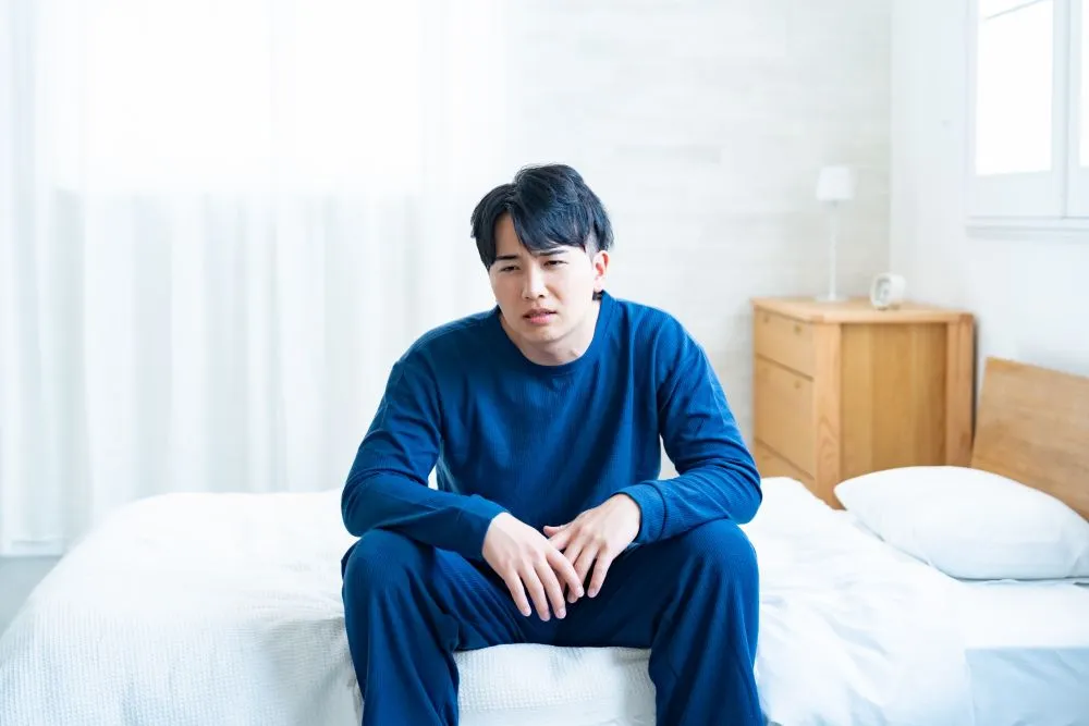 青い上下のパジャマを着た男性が白い寝具に座り、眠そうでつらい表情をしながら前を見ている。白い壁と窓から光が差し込み、早朝覚醒のイメージを表す。