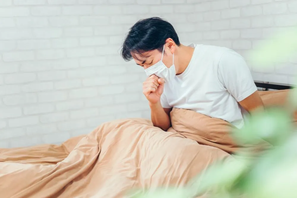 白いマスクと半袖Tシャツを着た若い男性が、ベージュ色の布団をかぶって座り込み、右手の拳で口元を押さえて激しく咳き込んでいる様子。夜更かしや睡眠不足で悪化した咳をイメージしたシーン。
