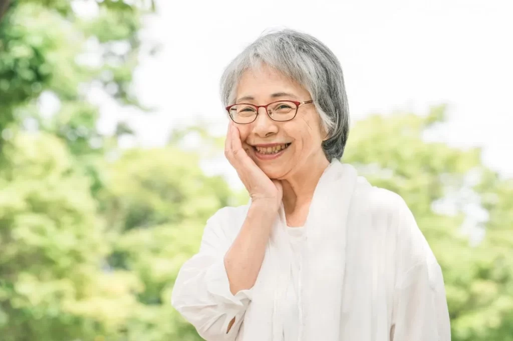 80代の高齢女性が新緑の公園で笑顔でほっぺを触っている。肝斑が鍼治療で改善した明るい表情