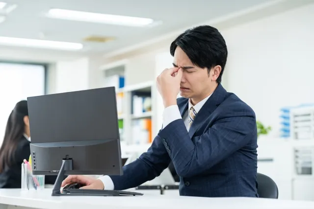 パソコン作業で目が疲れ、両目の間を押さえる男性。長時間前傾姿勢が後頭部神経痛の原因になる様子