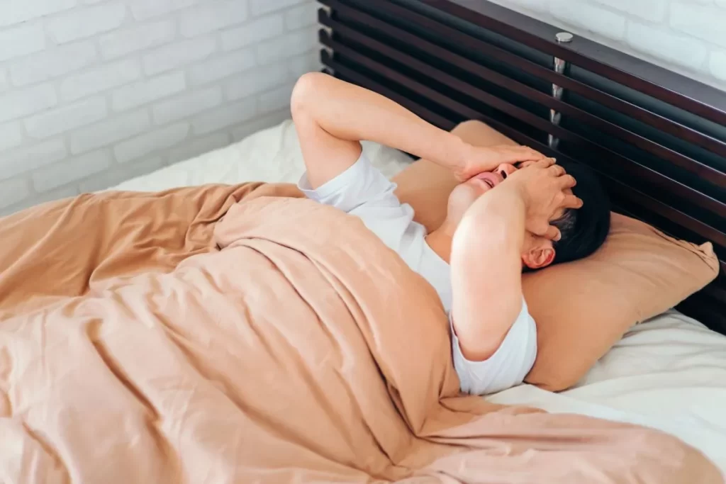 だるそうにベッドに寝て、両手で頭を触る白い半袖の男性