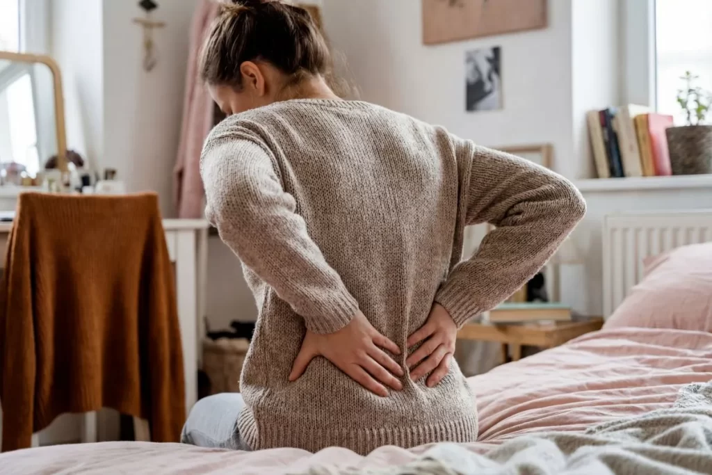 背中の痛みでつらそうにベッドに座り、両手で背中を押さえる女性の後ろ姿