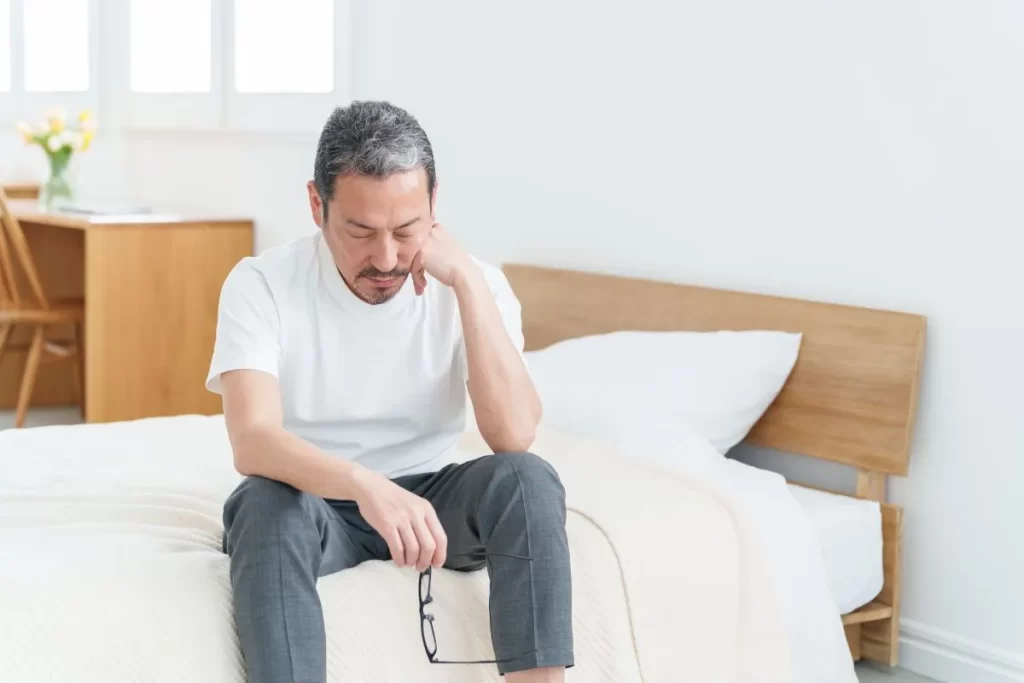 疲れた表情でベッドに座り、メガネを手に持ちながら目を閉じて休む中高生男性の写真