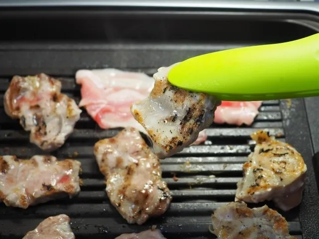 緑のトングで焼いた肉をつまんでいる写真。重い料理が脾胃に負担をかけ、食後の強い眠気につながる説明用。