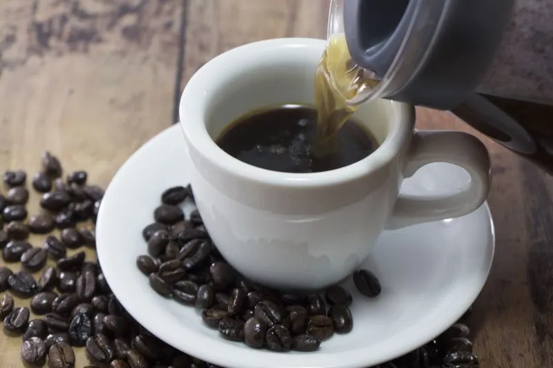 白いコップにコーヒーを注ぐ様子と木製テーブルに散らばったコーヒー豆の写真