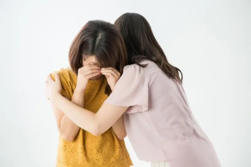 ベージュの服の女性が泣きそうな女性の肩を抱いて慰めている