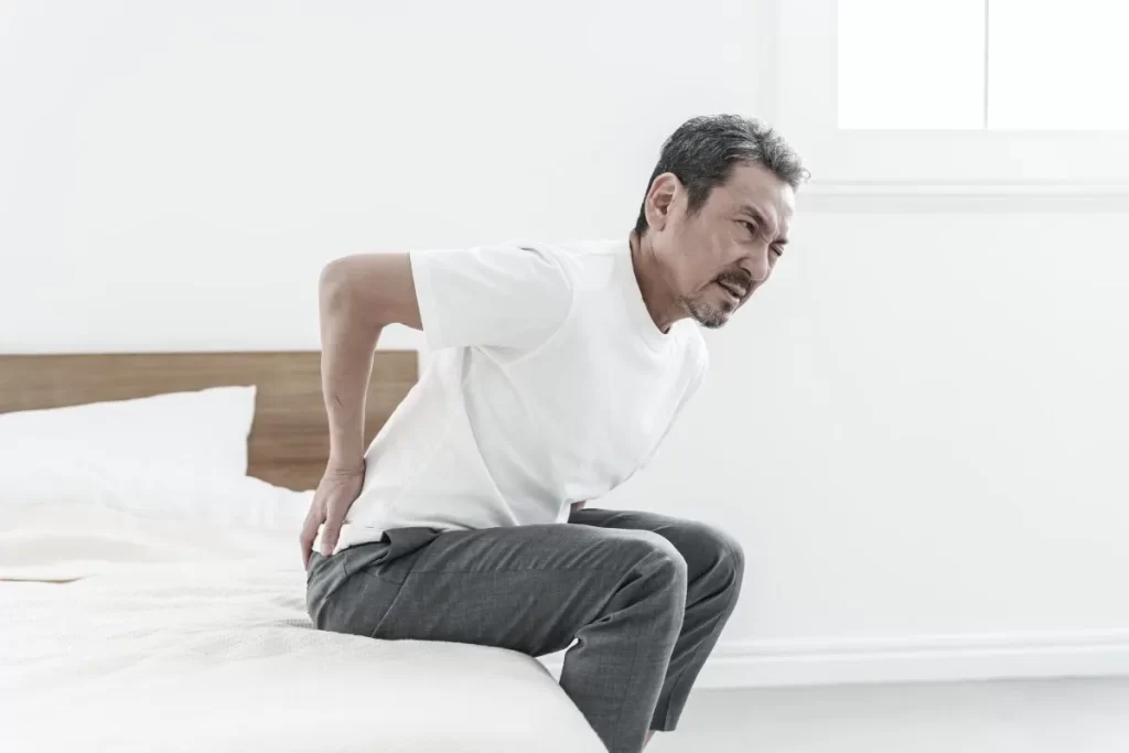 腰の重だるさに辛い表情を浮かべ、両手で腰を押さえながらベッドから起き上がろうとする中年男性